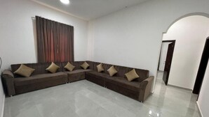 A spacious living room with a large L-shaped sectional sofa, a flat-screen TV, and a large window with curtains. - RED3 NEW SALALAH (Salalah)