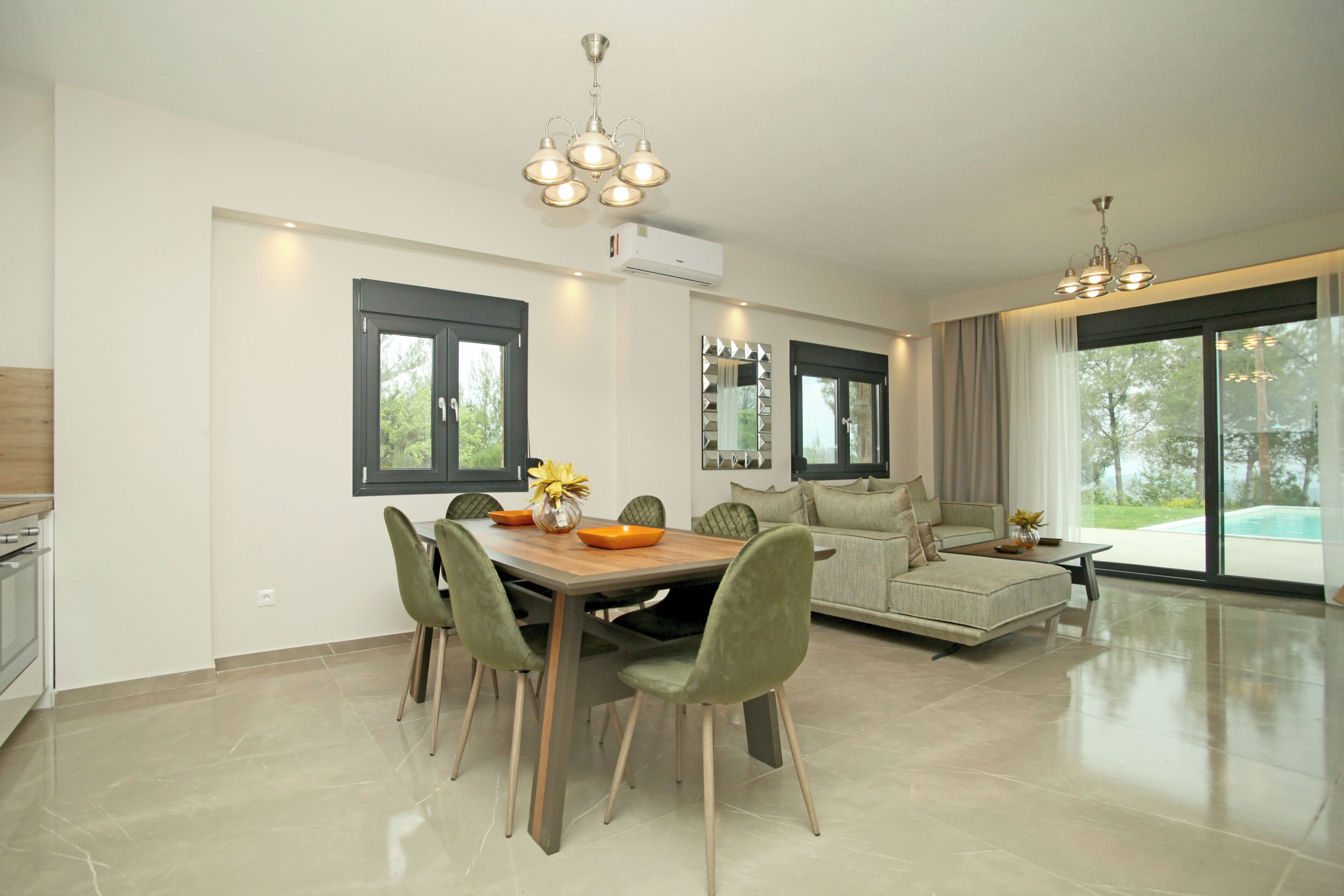 A modern dining area with a wooden table, green chairs, and a chandelier. There is a sofa and a large window with curtains.