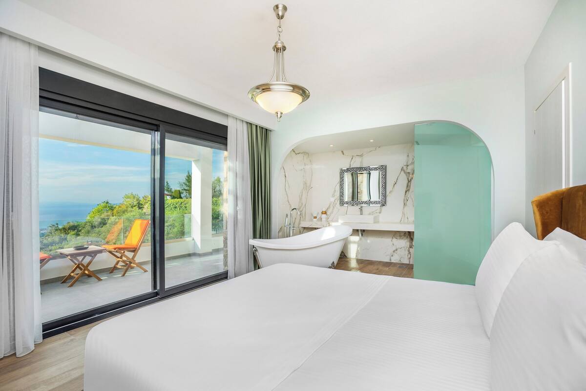 Ein modernes Hotelzimmer mit einem großen Bett, einer Badewanne, einem Balkon mit Stühlen und Blick auf das Meer.