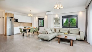 A modern kitchen with white cabinets, a stainless steel refrigerator, and a dining area with a green chair and a wooden table. - Epta Villas Luxury Redefined (Kassandra)