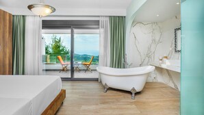 A modern bedroom with a large bed, a freestanding bathtub, and a balcony with chairs. - Epta Villas Luxury Redefined (Kassandra)