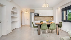 A modern kitchen with white cabinets, a wooden countertop, and a stainless steel refrigerator. There is a dining table with four chairs and a chandelier above. - Epta Villas Luxury Redefined (Kassandra)