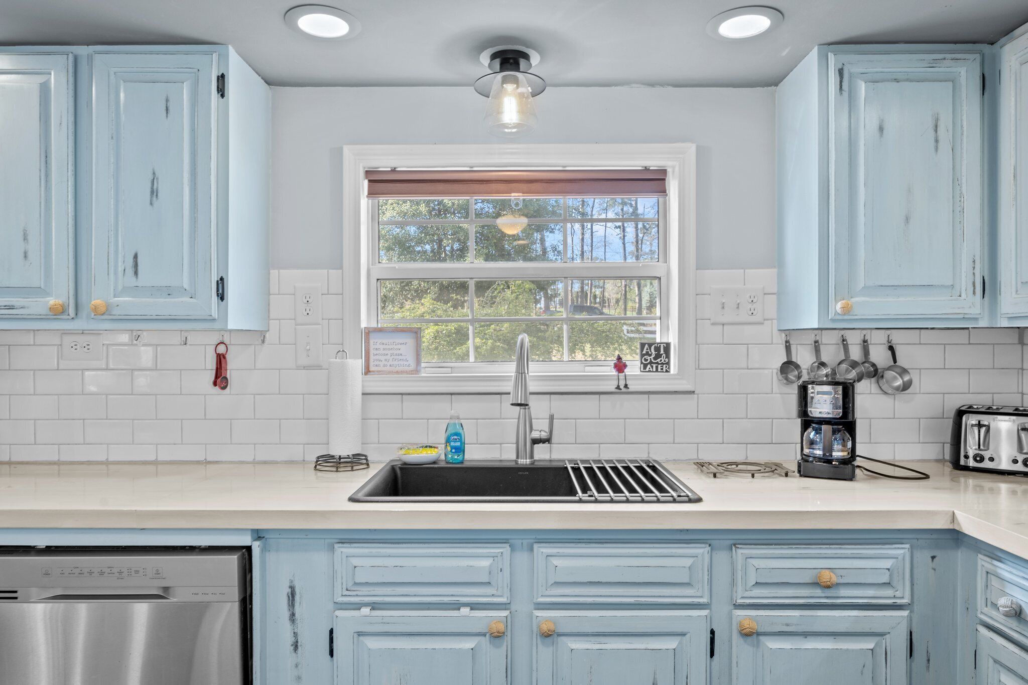 Una cocina con armarios azul claro, un lavavajillas de acero inoxidable, un fregadero con un grifo moderno y una ventana con vista a árboles.