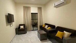 A modern living room with a marble floor, a dark sofa set with yellow cushions, a small coffee table, a TV mounted on the wall, and an air conditioning unit. - RED-33 - Gardenia (Salalah)