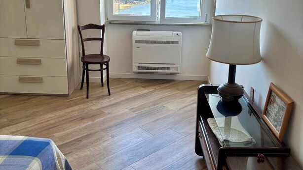 A room with a wooden floor, a bed with a blue and white checkered blanket, a chair, a table with a lamp, a picture frame, and a window with a view of the sea.