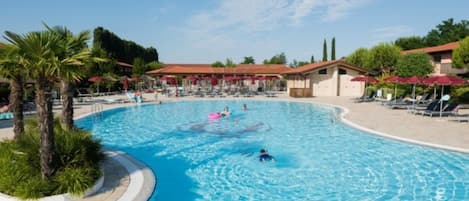 A large outdoor swimming pool with lounge chairs and umbrellas, surrounded by palm trees and a building with a red roof.