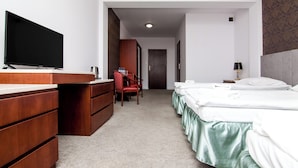 Camera d'albergo con una cassettiera in legno, TV a schermo piatto, una scrivania, una sedia, un letto con biancheria verde e un comodino con una lampada.