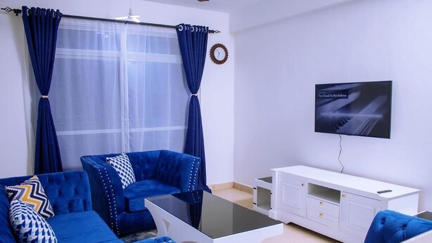 Ein Wohnzimmer mit blauen Sofas, einem Glastisch, einer weißen TV-Konsole und einem Fenster mit blauen Vorhängen.