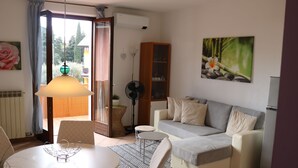 A living room with a sofa, a dining table with chairs, a fan, a bookshelf, and a window with curtains.