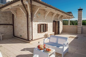 A covered outdoor seating area with stone walls, a white wicker sofa, and a table set with food and drinks. - Olive Grove (Puglia)