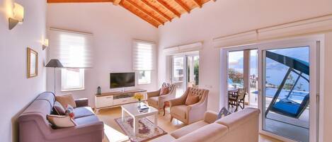 A spacious living room with a wooden ceiling, large windows, and a view of the sea.