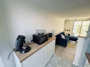 A compact kitchen with a coffee maker, microwave, and basket on a wooden countertop. A dark sofa and a small table are in the background.