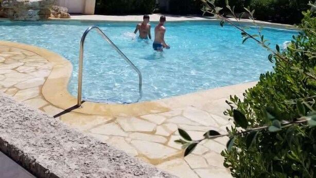 A swimming pool with two people in the water, surrounded by palm trees and a building with a tiled roof. - Valras-Loc – Petite maison avec jardin et piscine (Valras-Plage)