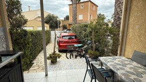A covered patio with a table and chairs, a red car parked outside, and a view of a residential area with buildings and trees. - Valras-Loc – Petite maison avec jardin et piscine (Valras-Plage)