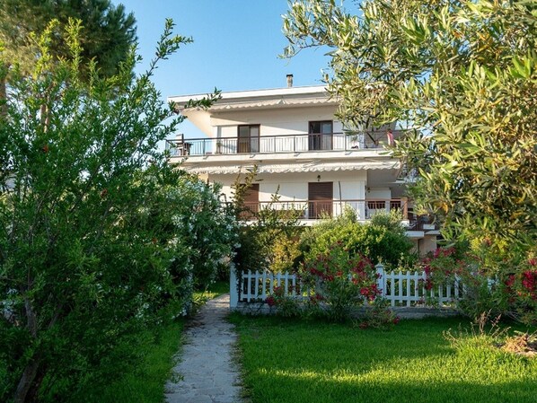 Un bâtiment à deux étages avec une façade blanche, un balcon à l’étage supérieur et une clôture blanche en bois devant la maison.