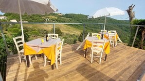 An outdoor dining area with tables and chairs, covered by umbrellas, overlooking a scenic hillside.