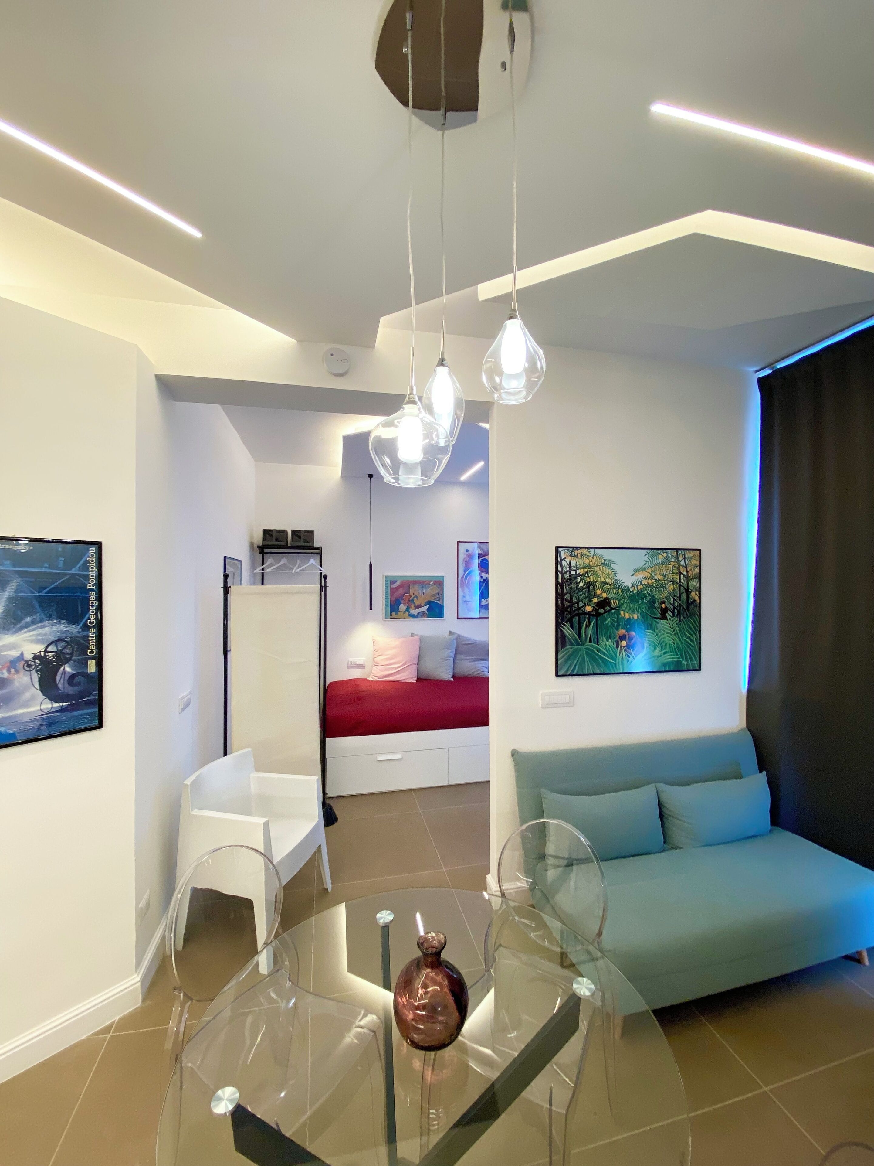 A modern living room with a glass coffee table, a sofa, a bed with a red blanket, and artwork on the walls.