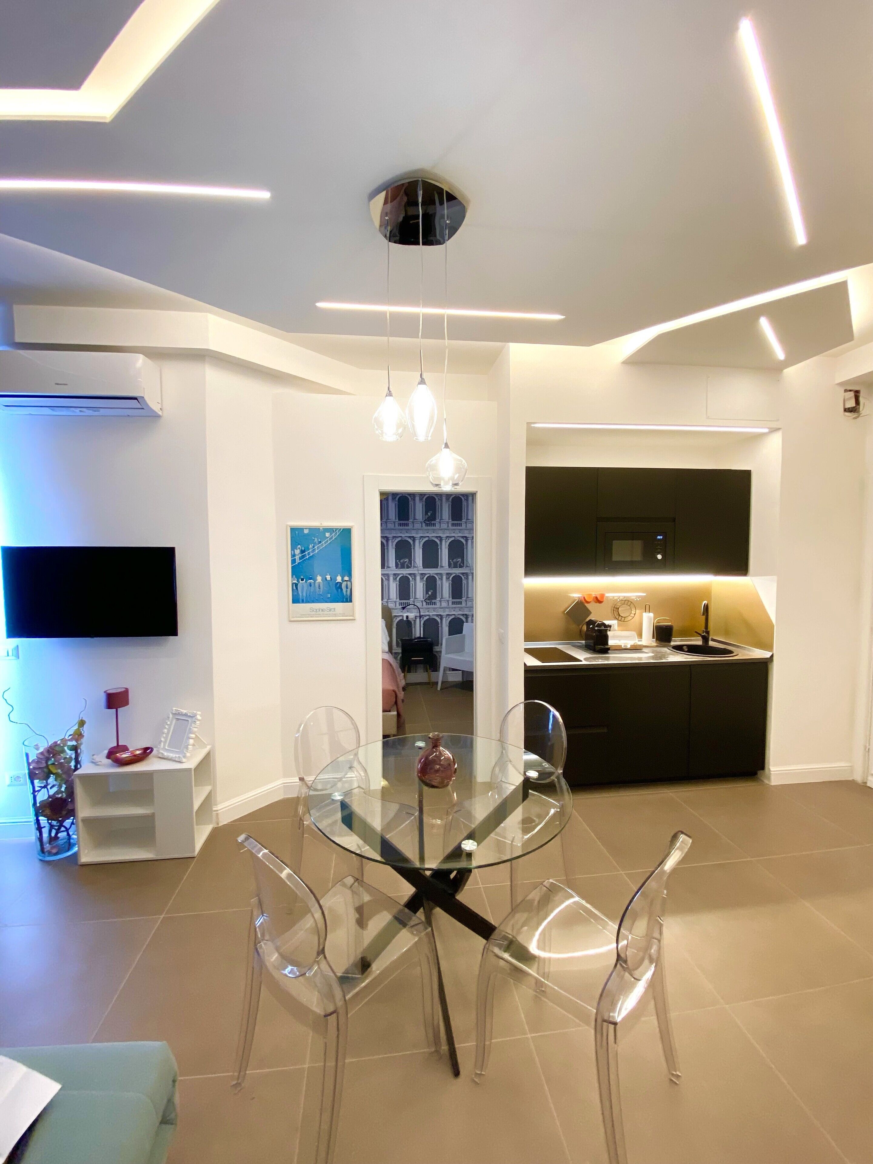 A modern living room with a glass dining table, clear chairs, a mounted TV, and a kitchen area with dark cabinetry.
