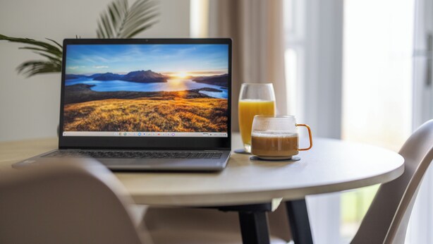 Un ordinateur portable posé sur une table, avec un papier peint représentant un paysage, un verre de jus d’orange et une tasse de café.