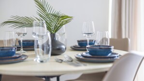 Une table à manger dressée avec des assiettes bleues, des verres transparents et un vase contenant une plante.