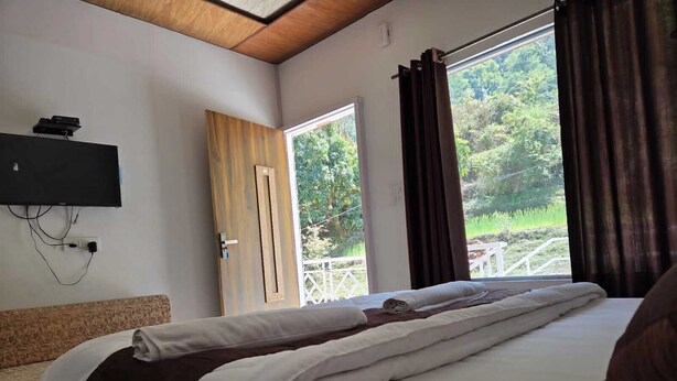 A bedroom with a bed, a TV mounted on the wall, and a window with curtains. - Namami Kedara Resort (Ukhimath)