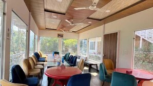 A modern indoor space with wooden ceiling beams, large windows, and a mix of round and square tables with colorful chairs. - Namami Kedara Resort (Ukhimath)