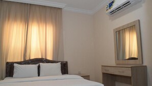 A hotel room with a bed, a wooden desk, a mirror, and an air conditioning unit.