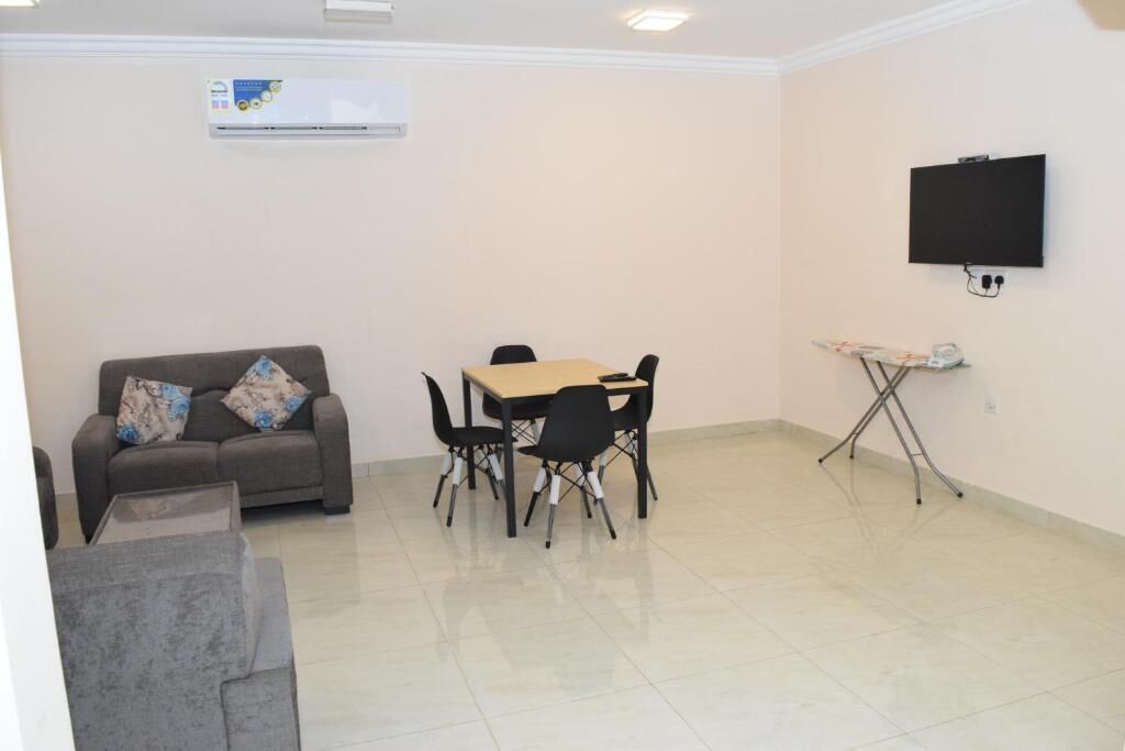 A room with a sofa, a dining table with chairs, a flat-screen TV mounted on the wall, and an ironing board.