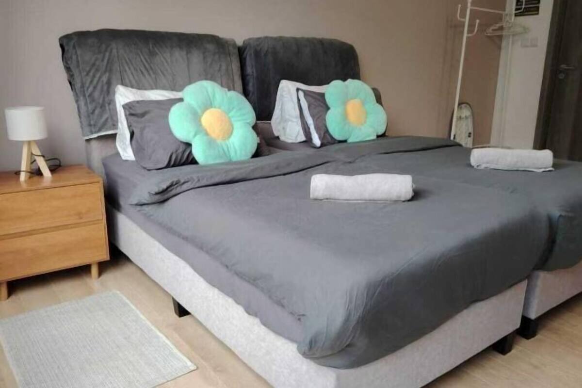 A bed with grey bedding, two flower-shaped pillows, a white towel, a wooden nightstand with a lamp, and a grey rug.