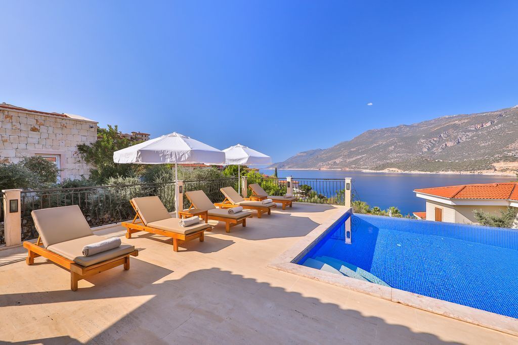 A poolside area with lounge chairs and umbrellas, overlooking a body of water and hills.