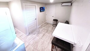 Ein kleines Zimmer mit weißer Tür, einem Tisch und einer Bank, einem an der Wand befestigten Fernseher sowie einer Decken-Klimaanlage.