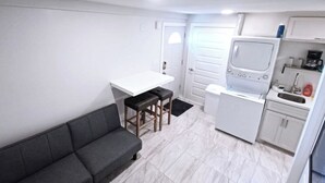 Eine kompakte Küche mit weißer Arbeitsplatte, einem kleinen Kühlschrank und einer Mikrowelle. Im Wohnbereich befindet sich eine graue Couch und zwei Hocker.