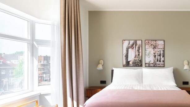 Un dormitorio con una cama grande, un banco, una ventana con cortinas y dos lámparas fijadas a la pared.