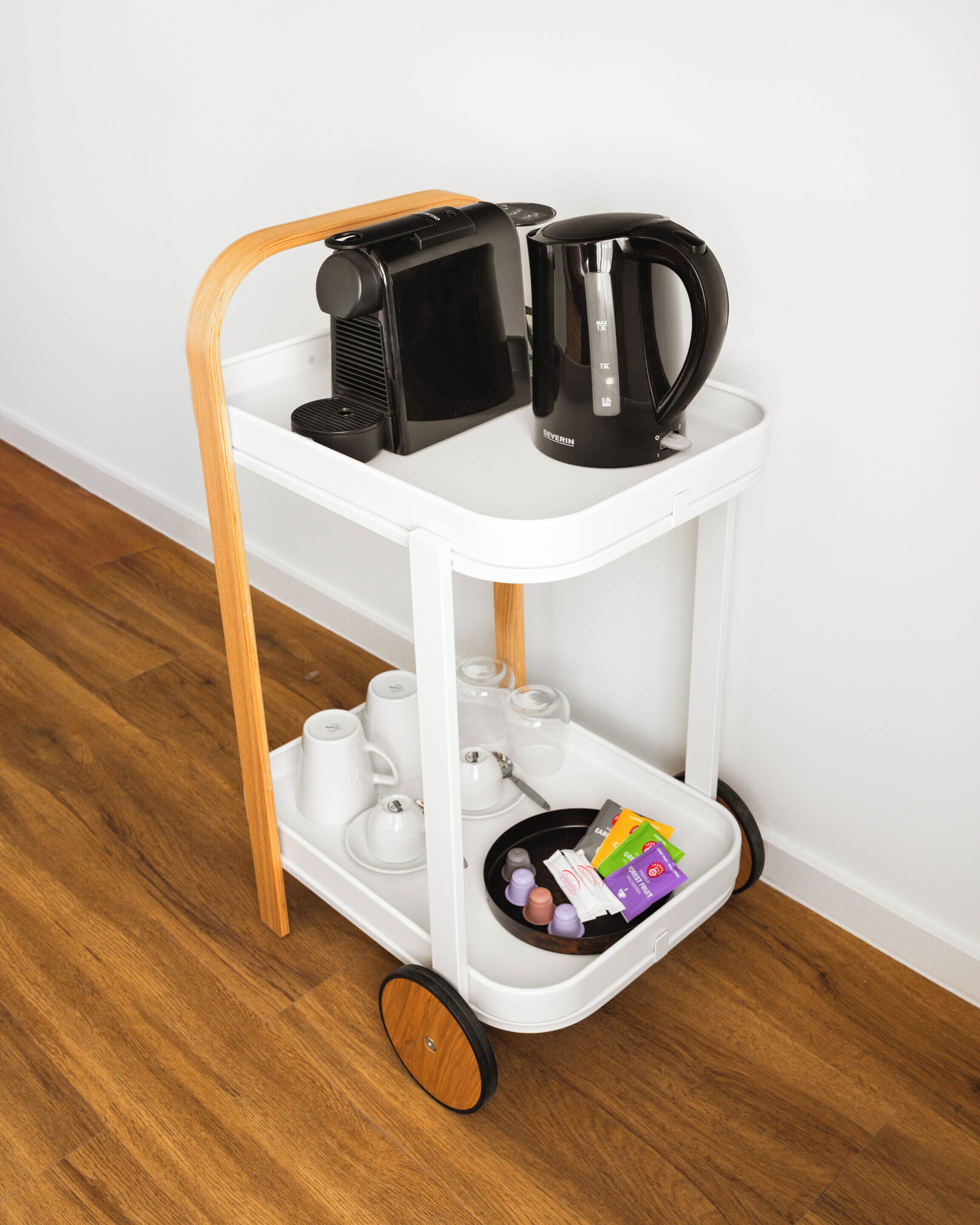 A white mobile cart with a wooden handle, containing a black coffee maker, a black electric kettle, white cups, and a tray with small items.