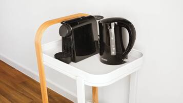 A white mobile cart with a wooden handle, containing a black coffee maker, a black electric kettle, white cups, and a tray with small items.
