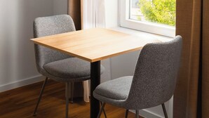Una habitación moderna con una mesa de madera y dos sillas grises, una ventana con cortinas y vistas a edificios y árboles.