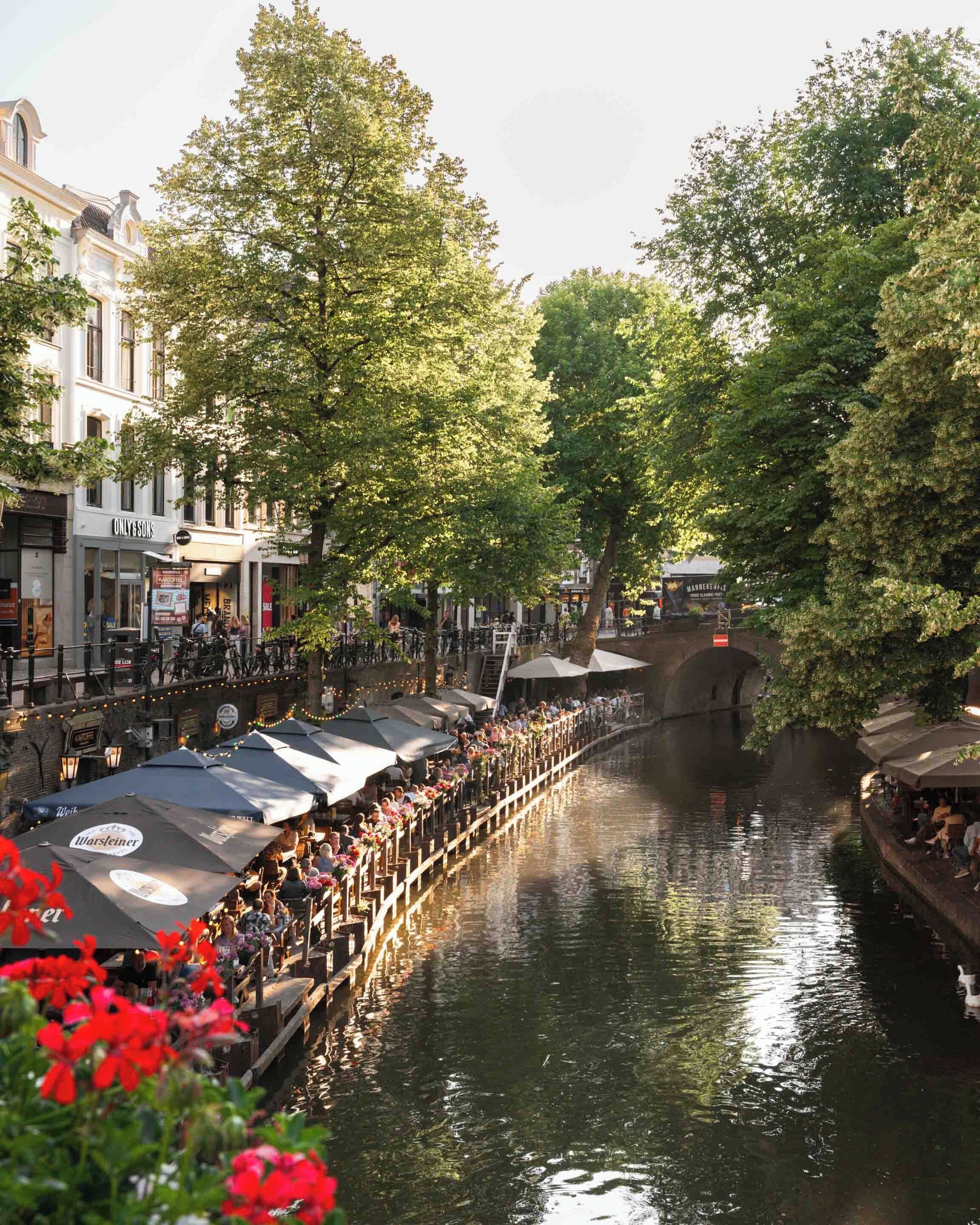 A riverside promenade with outdoor seating areas, a bridge, and buildings along the water.