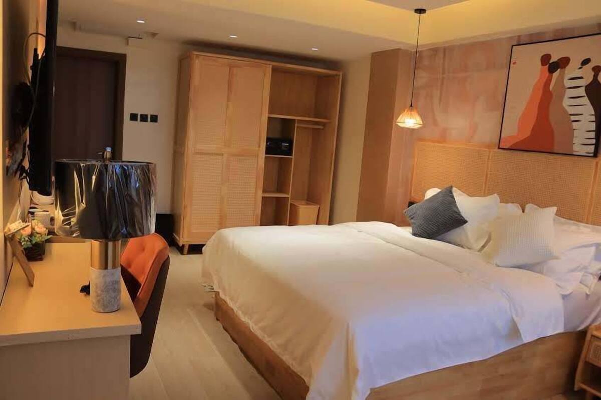 A hotel room with a bed, a desk with a lamp, a wardrobe, and a painting on the wall.