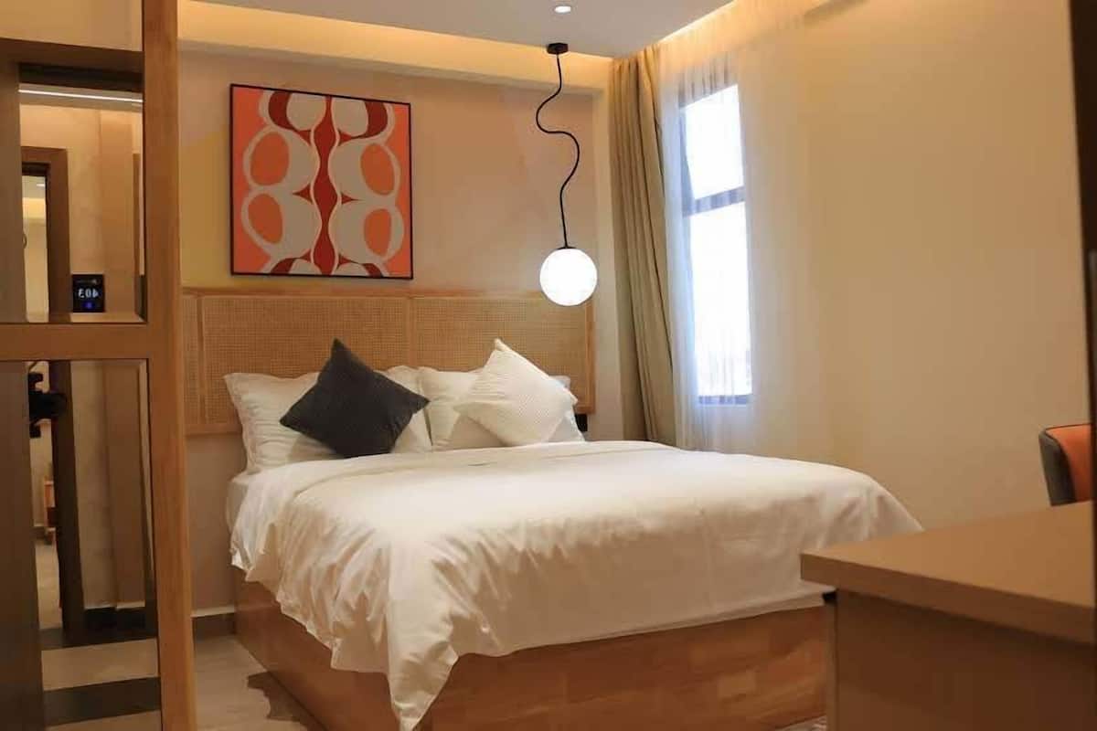 A hotel room with a bed, a desk, a wall-mounted artwork, and a hanging light fixture.