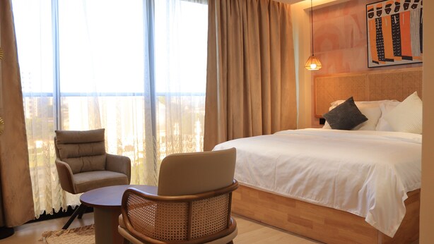 A hotel room with a bed, two chairs, a table, a large window with curtains, and a wall-mounted artwork. - Arova Hotel (Nairobi)