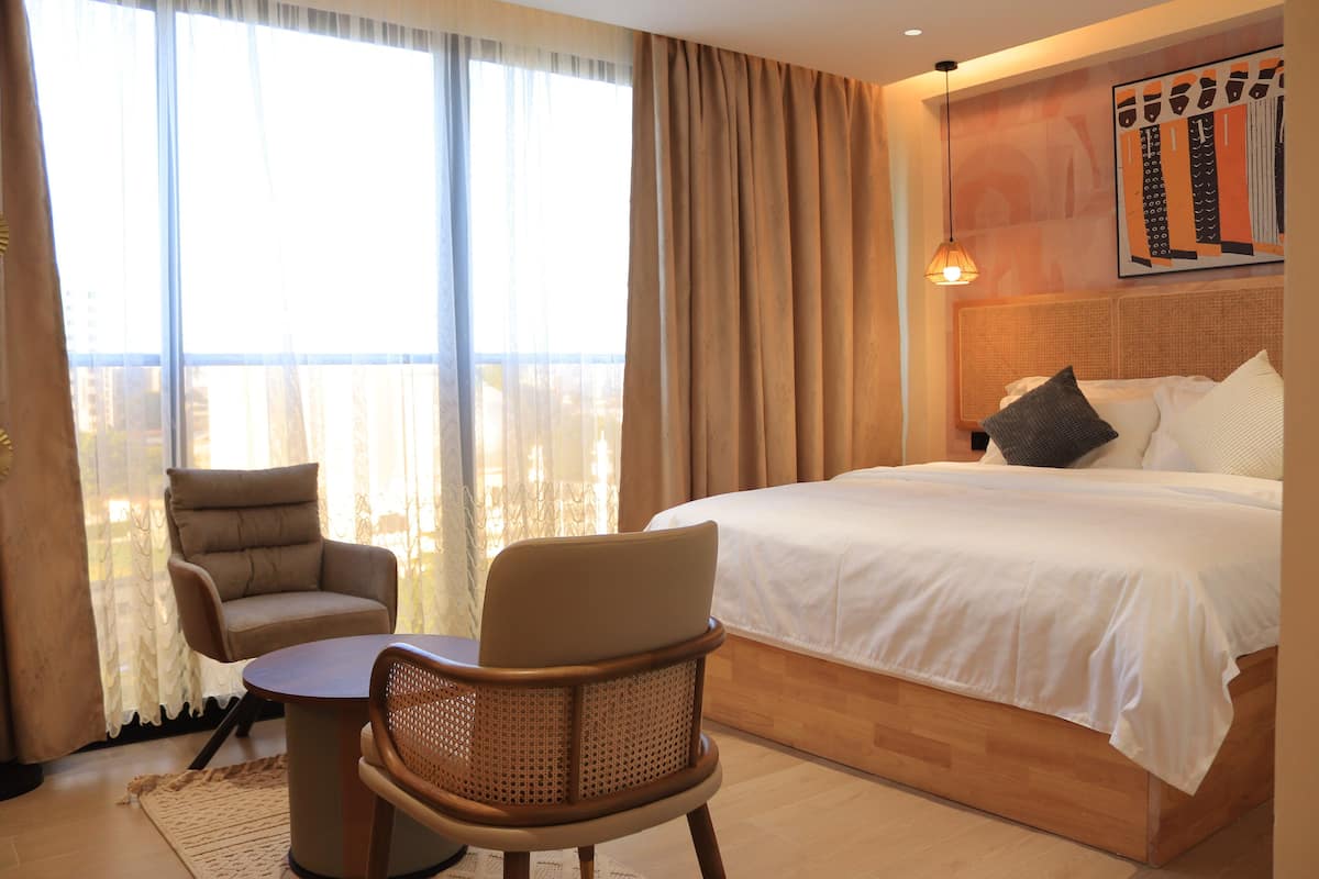 A hotel room with a bed, two chairs, a table, a large window with curtains, and a wall-mounted artwork.