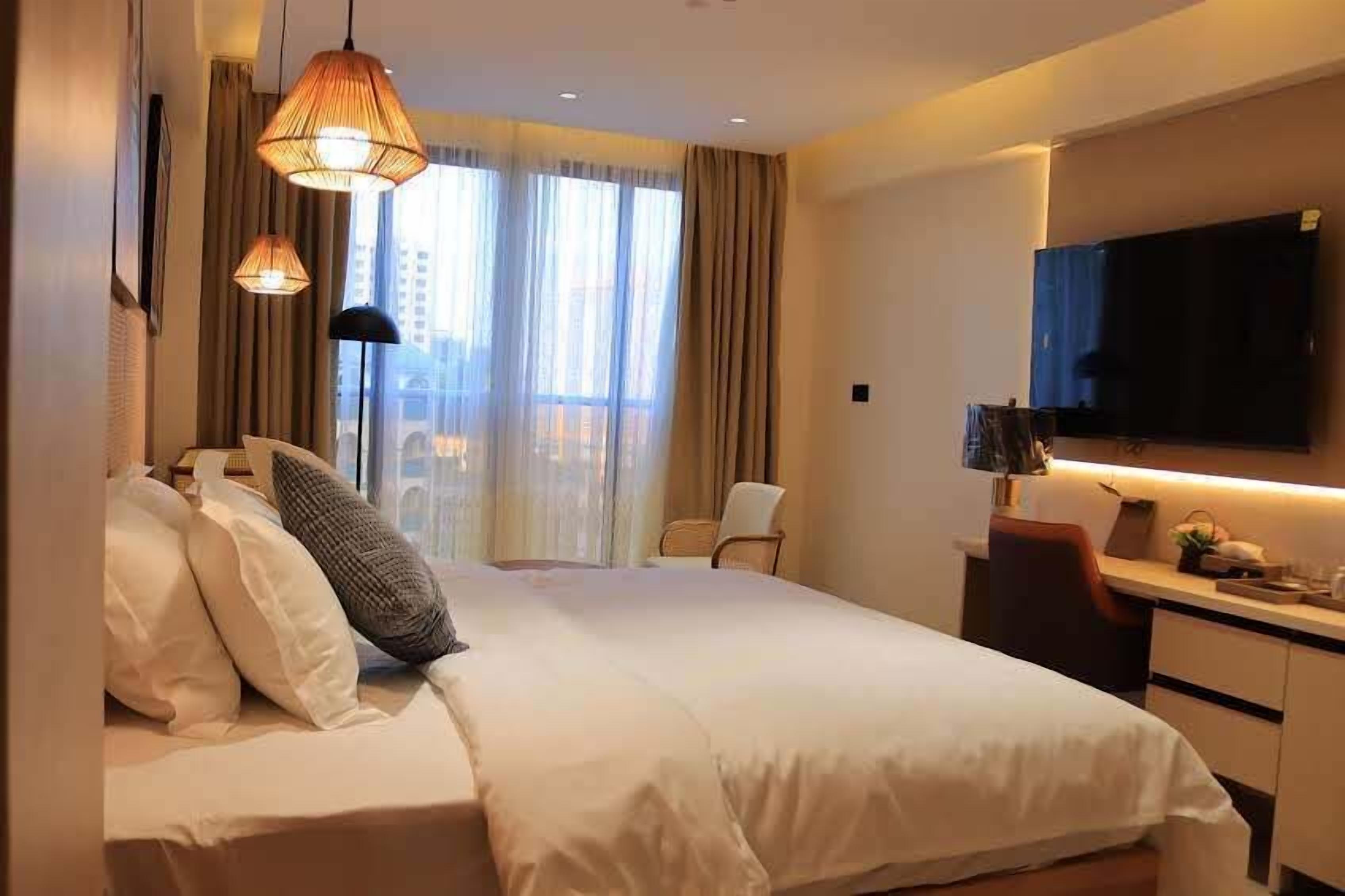 A hotel room with a large bed, a desk with a flat-screen TV, and a view of the city through large windows.