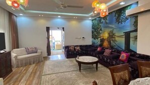 A spacious living room with a large sofa, a coffee table, and a dining area with chairs. There is a ceiling fan, a chandelier, and a painting on the wall. - Red 12 Almasaraat (Salalah)