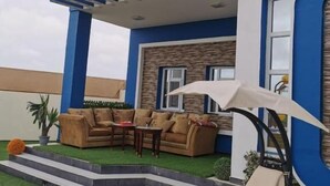 A modern house with a blue and white exterior, a covered patio with a sofa and chairs, and a small garden area with artificial grass and a lounge chair. - Red 12 Almasaraat (Salalah)