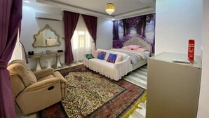 A bedroom with a bed, a massage chair, a vanity with a mirror, and a large window with curtains. - Red 12 Almasaraat (Salalah)