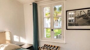 Ein Schlafzimmer mit einem Bett, einem Fenster mit Vorhängen, einer kleinen Bank und einem gerahmten Bild an der Wand.