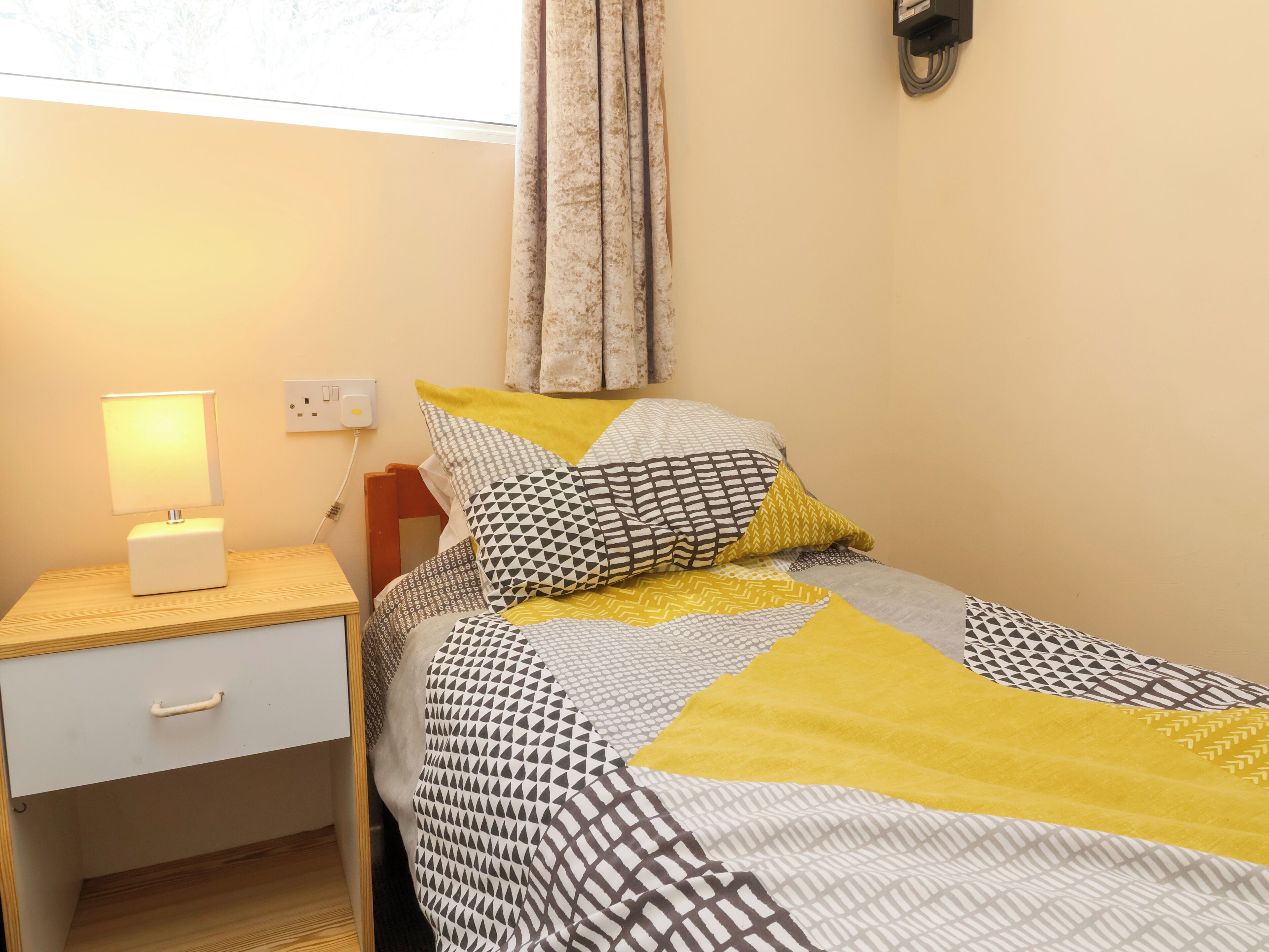 A neatly made bed with patterned bedding, a bedside table with a lamp, and a power outlet on the wall.