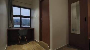 A small, dimly lit room with a wooden desk and chair, a large window, and a door with a mirror.