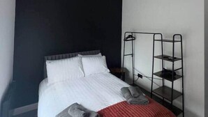 Hotellihuone, jossa on sänky, punainen peitto, valkoinen tyyny, musta seinä, metallinen hylly ja kattovalaisin.