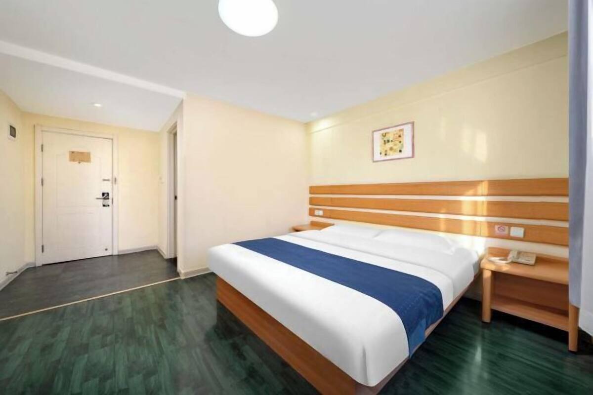 A hotel room with a bed, a bedside table with a phone, a door, and a wall-mounted picture.